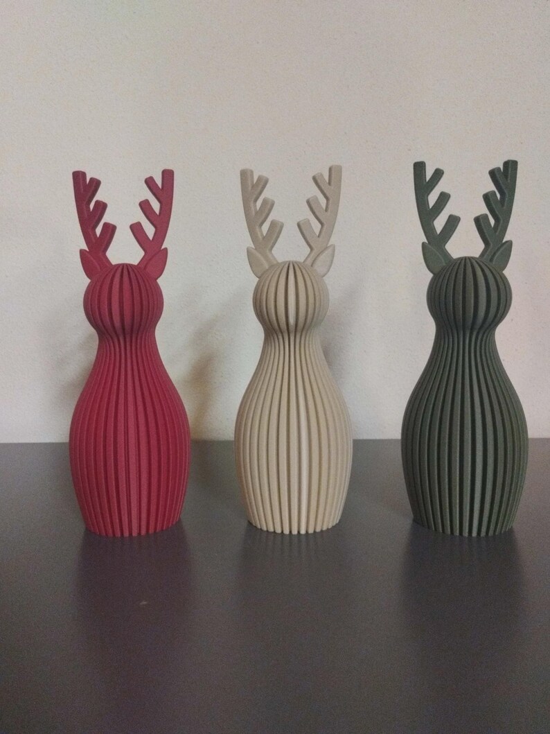 May include: Three standing reindeer figurines with a ribbed, vertical slat construction. The left figure is deep red, the center figure is off-white, and the right figure is dark olive green. Each has a rounded body and branched antlers.