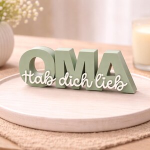 K&ouml;nnte beinhalten: Hellgr&uuml;ne Holzbuchstaben mit der Aufschrift "OMA" und den Worten "Hab dich lieb" in wei&szlig;er Schrift. Die Buchstaben stehen auf einem hellen Holztablett, mit einer Kerze und einer Blumenvase im Hintergrund.