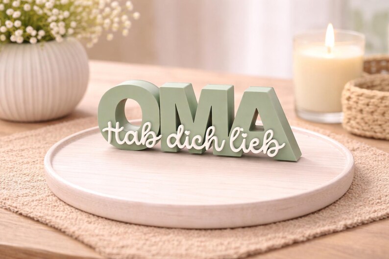 K&ouml;nnte beinhalten: Hellgr&uuml;ne Holzbuchstaben mit der Aufschrift "OMA" und den Worten "Hab dich lieb" in wei&szlig;er Schrift. Die Buchstaben stehen auf einem hellen Holztablett, mit einer Kerze und einer Blumenvase im Hintergrund.
