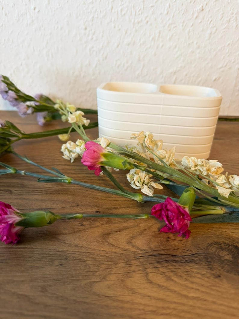 May include: A matte white ceramic toothbrush holder with a rectangular shape and horizontal ribbed detailing, accompanied by several stems of pink carnations and dried white flowers arranged in the foreground.