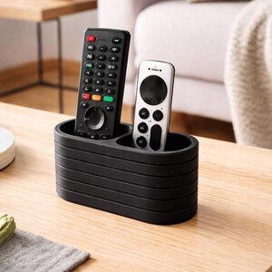 May include: A matte black, oval-shaped remote control holder with horizontal ribbed detailing. The organizer features two distinct compartments, currently holding one tall black remote and one shorter silver remote.