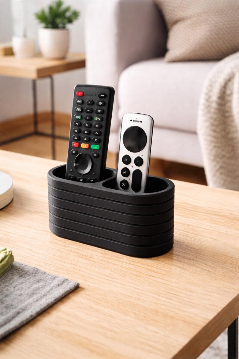 May include: A matte black, oval-shaped remote control holder with horizontal ribbed detailing. The organizer features two distinct compartments, currently holding one tall black remote and one shorter silver remote.