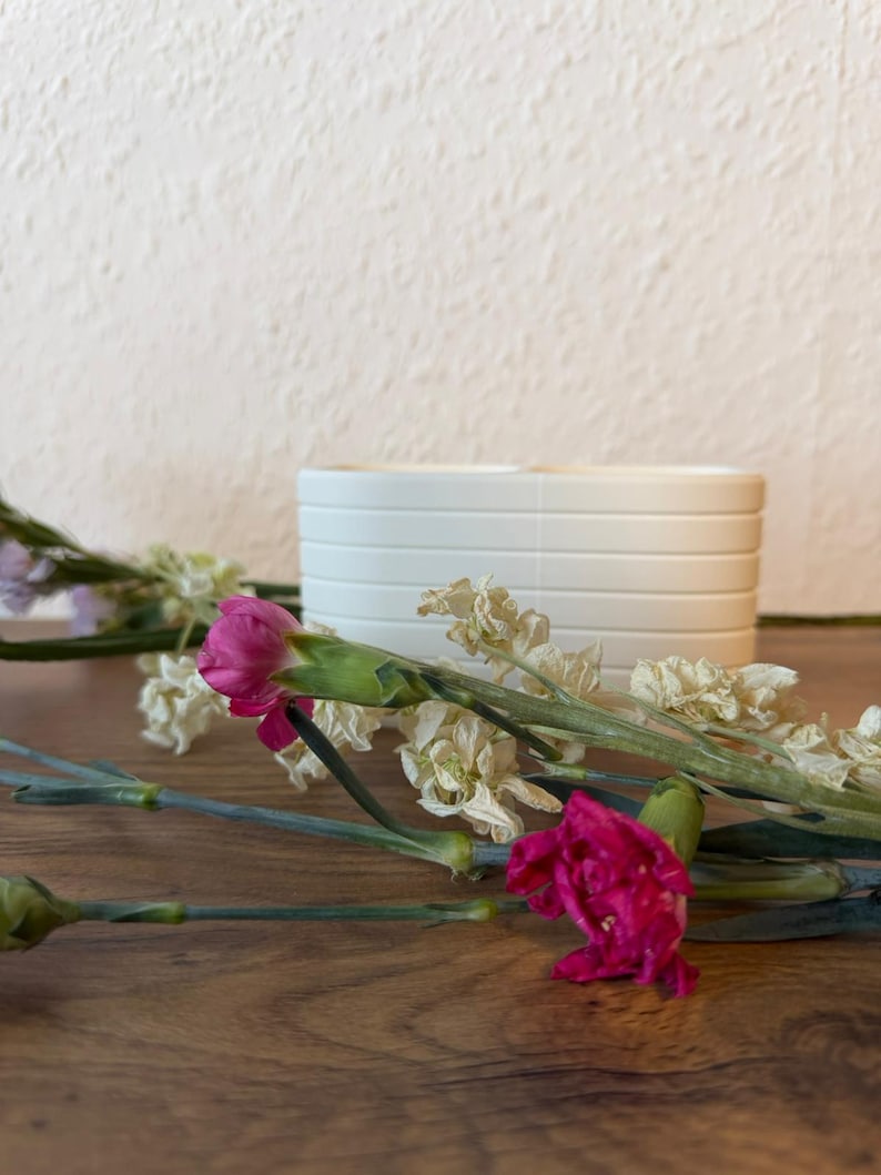 May include: A white ceramic planter with horizontal ribbed detailing, positioned behind several stems of vibrant magenta carnations and dried white flowers, showcasing a clean and structured design for plant display.