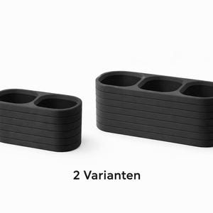 May include: Two matte black organizers with rounded rectangular shapes. The smaller organizer features two circular compartments, while the larger one has three. Both items display horizontal ribbed detailing and the text 2 Varianten.