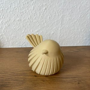May include: A small, light beige bird figurine with a rounded body and a textured, ribbed surface pattern. The bird features a pointed beak and a fanned tail, showcasing a matte finish with visible 3D printed layer lines throughout the structure.