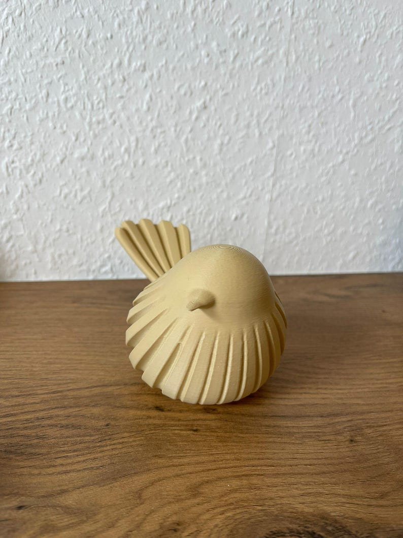 May include: A small, light beige bird figurine with a rounded body and a textured, ribbed surface pattern. The bird features a pointed beak and a fanned tail, showcasing a matte finish with visible 3D printed layer lines throughout the structure.