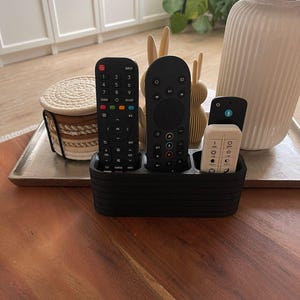 May include: A black, rectangular remote control holder with horizontal ribbed detailing, featuring three distinct compartments. It contains a long black remote with a numeric keypad, a taller black remote with a circular navigation pad, and a smaller white remote.