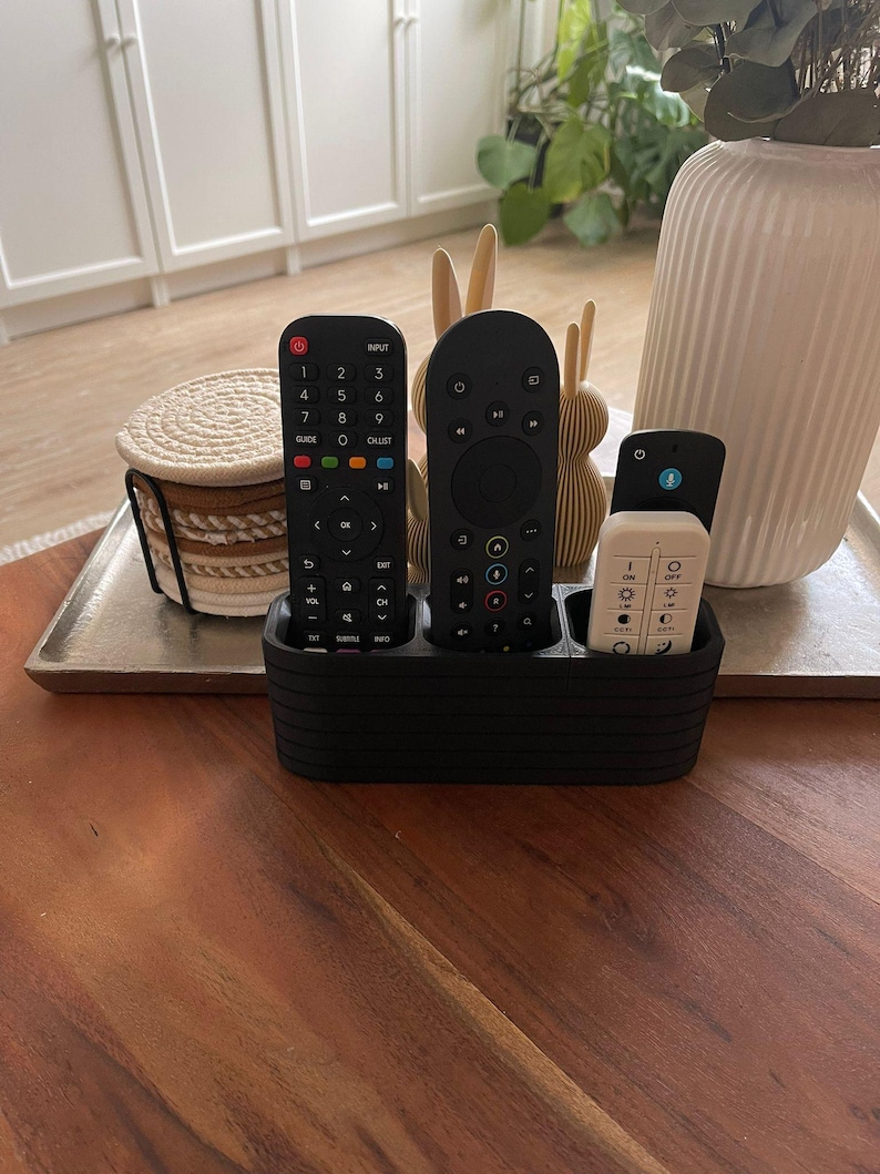 May include: A black, rectangular remote control holder with horizontal ribbed detailing, featuring three distinct compartments. It contains a long black remote with a numeric keypad, a taller black remote with a circular navigation pad, and a smaller white remote.