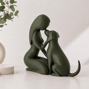 May include: A dark olive green sculpture depicting a stylized figure kneeling and touching foreheads with a dog. The smooth, matte-finished piece features minimalist, flowing lines that define the forms of both the human-like figure and the canine.