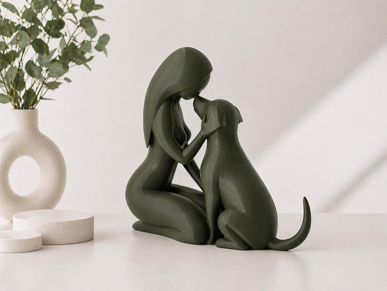 May include: A dark olive green sculpture depicting a stylized figure kneeling and touching foreheads with a dog. The smooth, matte-finished piece features minimalist, flowing lines that define the forms of both the human-like figure and the canine.