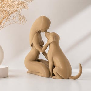 May include: A smooth, tan-colored figurine depicting a stylized human figure kneeling and touching foreheads with a dog. The sculpture features minimalist, flowing lines and a solid matte finish, capturing a moment of connection between the two figures.