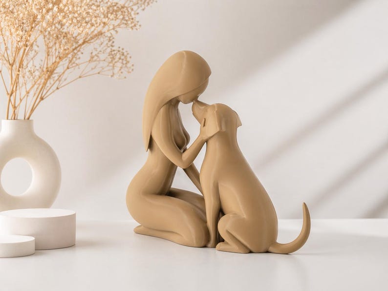 May include: A smooth, tan-colored figurine depicting a stylized human figure kneeling and touching foreheads with a dog. The sculpture features minimalist, flowing lines and a solid matte finish, capturing a moment of connection between the two figures.