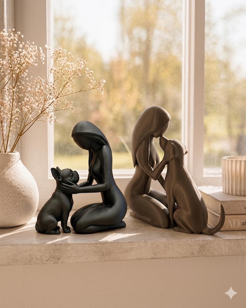 May include: Two smooth, matte-finish sculptures depicting figures kneeling and interacting with dogs. One sculpture is black, showing a figure with a French Bulldog, and the other is a dark gray, showing a figure kissing a dog on the nose.