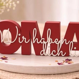 K&ouml;nnte beinhalten: Ein rotes Holzschild mit der Aufschrift "OMA" und den Worten "Wir haben dich lieb" in wei&szlig;er Schrift. Das Schild steht auf einem wei&szlig;en Teller, mit Makronen und einer brennenden Kerze im Hintergrund. Eine Geschenkbox und Blumen sind ebenfalls zu sehen.