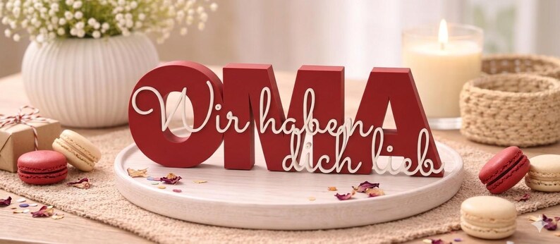 K&ouml;nnte beinhalten: Ein rotes Holzschild mit der Aufschrift "OMA" und den Worten "Wir haben dich lieb" in wei&szlig;er Schrift. Das Schild steht auf einem wei&szlig;en Teller, mit Makronen und einer brennenden Kerze im Hintergrund. Eine Geschenkbox und Blumen sind ebenfalls zu sehen.