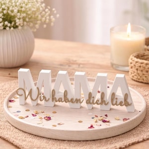 K&ouml;nnte beinhalten: Wei&szlig;e Holzbuchstaben mit der Aufschrift "MAMA" und goldfarbenem Schriftzug "Wir haben dich lieb" auf einem Holztablett. Das Tablett ist mit Konfetti und getrockneten Blumen verziert. Eine Kerze und eine Blumenvase befinden sich im Hintergrund.
