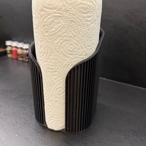 May include: A black paper towel holder with a vertical ribbed texture, featuring a wide front opening that displays a roll of white paper towels with a repeating embossed swirl pattern throughout the surface of the paper.