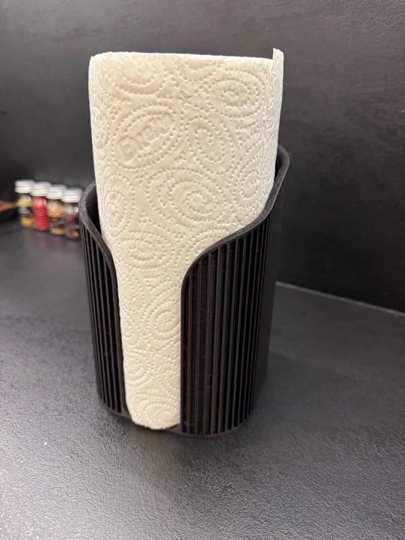May include: A black paper towel holder with a vertical ribbed texture, featuring a wide front opening that displays a roll of white paper towels with a repeating embossed swirl pattern throughout the surface of the paper.