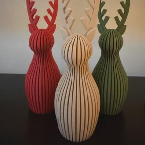 May include: Three standing reindeer figurines with a vertical ribbed texture. The left figure is red, the center figure is off-white, and the right figure is dark green. Each reindeer has a rounded body and branched antlers.