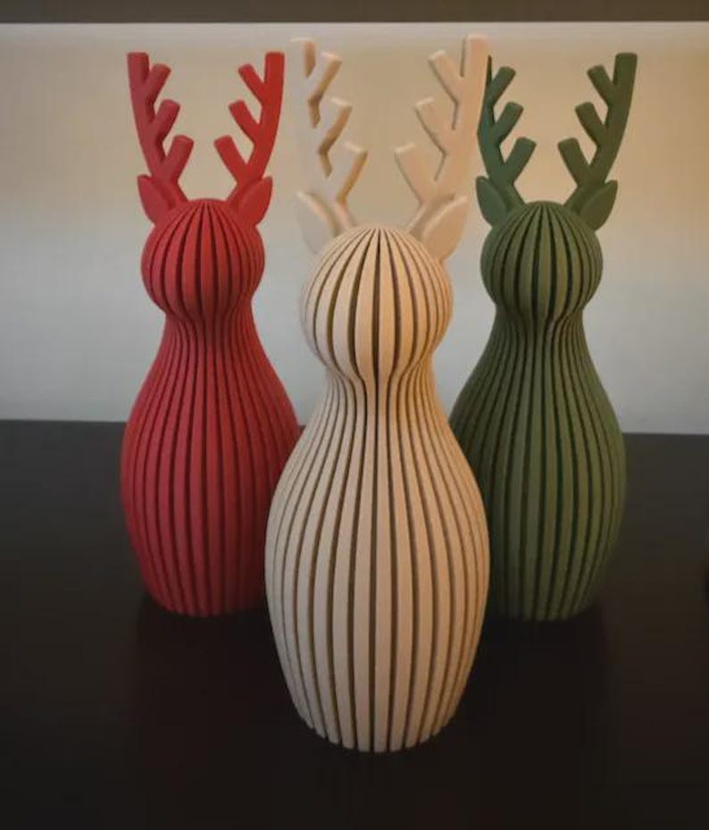 May include: Three standing reindeer figurines with a vertical ribbed texture. The left figure is red, the center figure is off-white, and the right figure is dark green. Each reindeer has a rounded body and branched antlers.