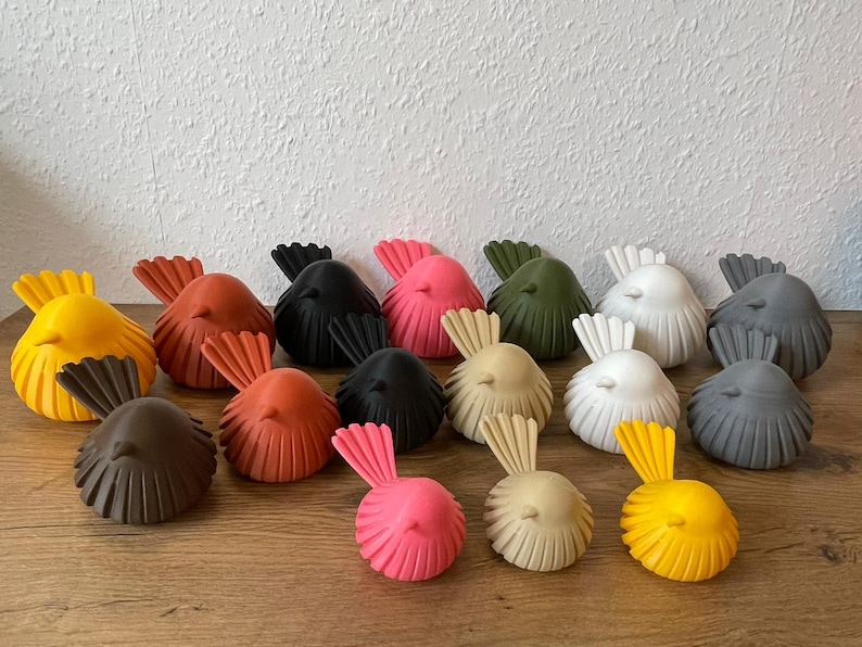 May include: A collection of fifteen small bird figurines in various solid colors including yellow, orange, black, pink, olive green, white, gray, brown, and beige. Each bird features a rounded body with vertical ribbed detailing and a fan-shaped tail.