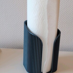 May include: A dark gray paper towel holder with a vertical ribbed texture. The holder features a curved, open-front design that securely contains a roll of white paper towels with a subtle embossed pattern on the surface of the paper.