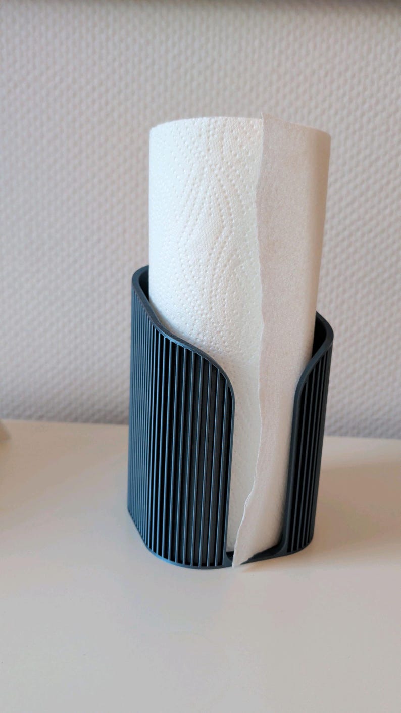 May include: A dark gray paper towel holder with a vertical ribbed texture. The holder features a curved, open-front design that securely contains a roll of white paper towels with a subtle embossed pattern on the surface of the paper.