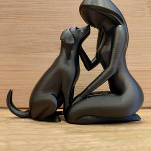 May include: A matte black figurine depicting a stylized human figure kneeling and interacting with a dog. The human figure has a smooth, featureless head and long hair, while the dog is shown sitting with its head tilted upward toward the human.