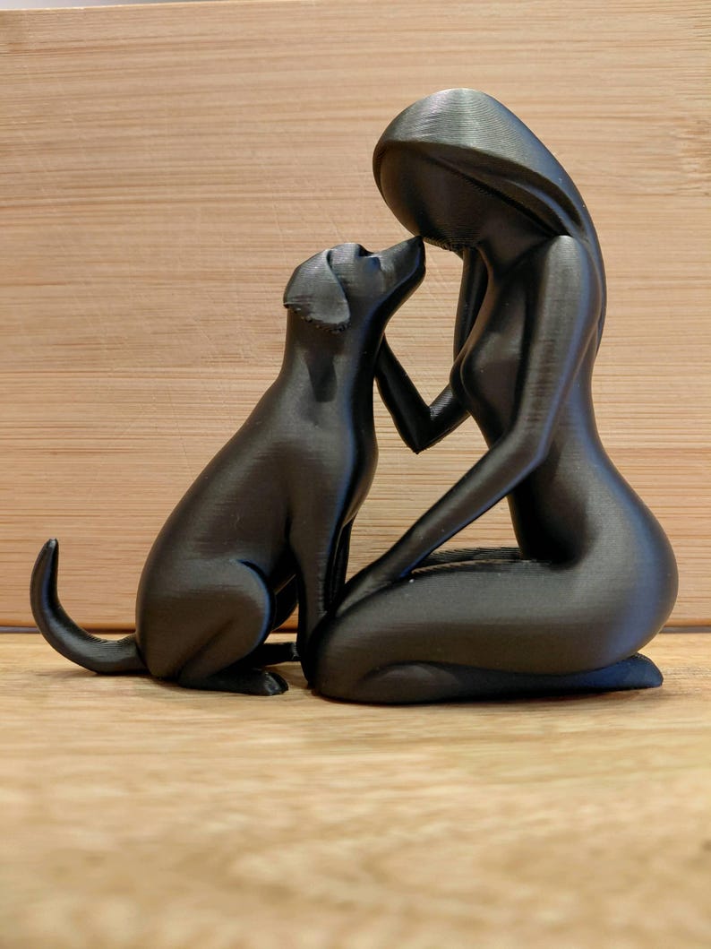 May include: A matte black figurine depicting a stylized human figure kneeling and interacting with a dog. The human figure has a smooth, featureless head and long hair, while the dog is shown sitting with its head tilted upward toward the human.