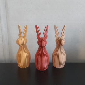 May include: Three standing reindeer figurines featuring a vertical ribbed texture. The left figure is yellow, the center figure is dark red, and the right figure is light tan. Each reindeer has a rounded body and branched antlers.
