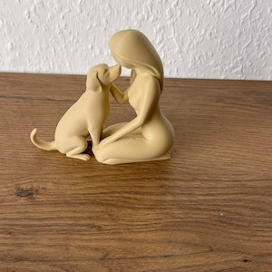 May include: A beige monochromatic figurine depicting a kneeling person with long hair facing a dog. The person is positioned on their knees with one hand reaching toward the dog's face, while the dog sits upright with its tail slightly curved.