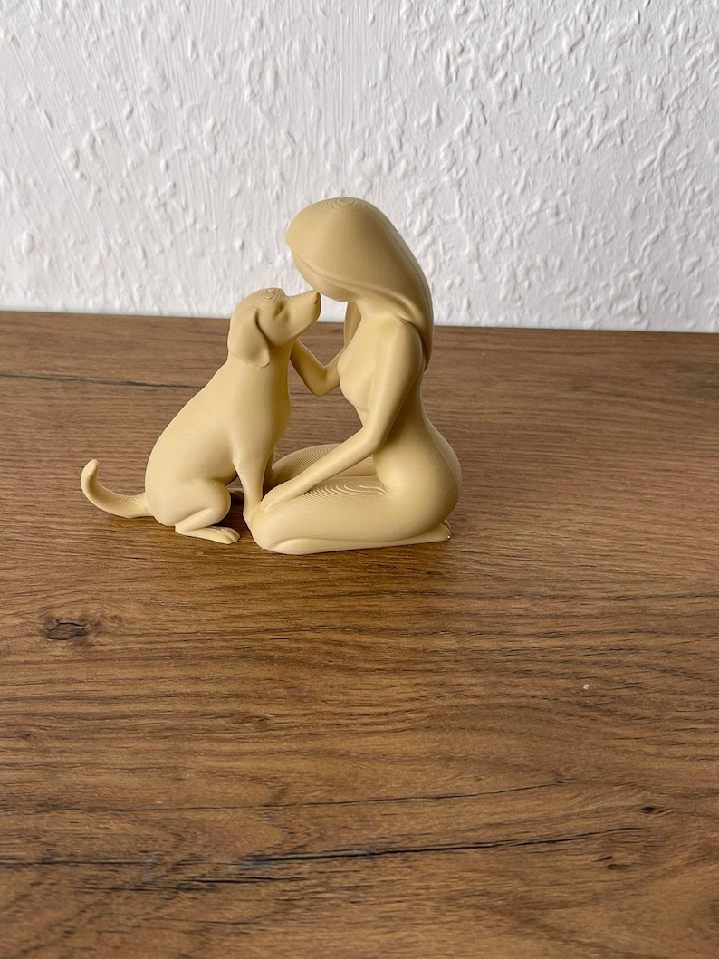 May include: A beige monochromatic figurine depicting a kneeling person with long hair facing a dog. The person is positioned on their knees with one hand reaching toward the dog's face, while the dog sits upright with its tail slightly curved.