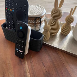 May include: A black plastic remote control organizer holding three remote controls, a stack of round white and tan rope coasters in a black metal holder, and three beige 3D-printed rabbit figurines on a rectangular silver tray.
