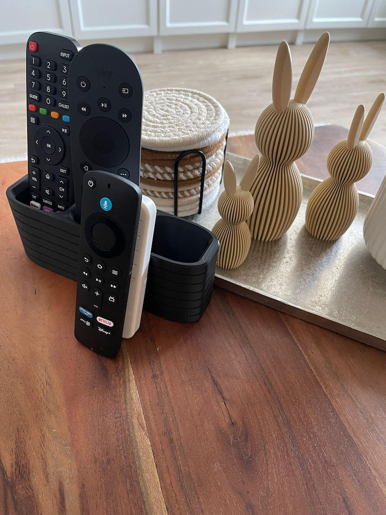 May include: A black plastic remote control organizer holding three remote controls, a stack of round white and tan rope coasters in a black metal holder, and three beige 3D-printed rabbit figurines on a rectangular silver tray.