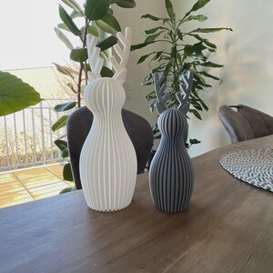 May include: Two standing deer figurines with a ribbed, vertical texture. The larger figurine is matte white, while the smaller figurine is matte gray. Both feature stylized antlers and a rounded, bulbous body shape with a flat base for stability.