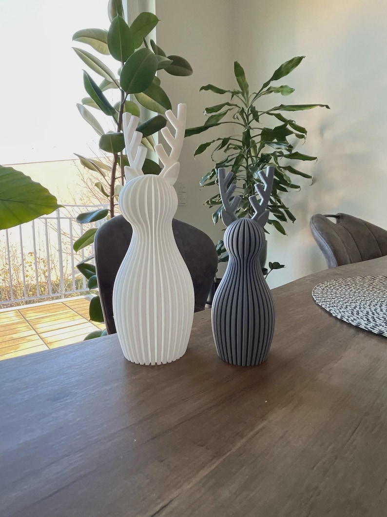May include: Two standing deer figurines with a ribbed, vertical texture. The larger figurine is matte white, while the smaller figurine is matte gray. Both feature stylized antlers and a rounded, bulbous body shape with a flat base for stability.