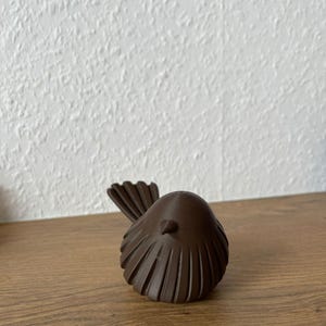 May include: A small, dark brown bird figurine with a rounded body and a textured, fan-shaped tail. The object features vertical ridges along its wings and a small, pointed beak, showcasing a smooth, matte finish throughout its entire structure.