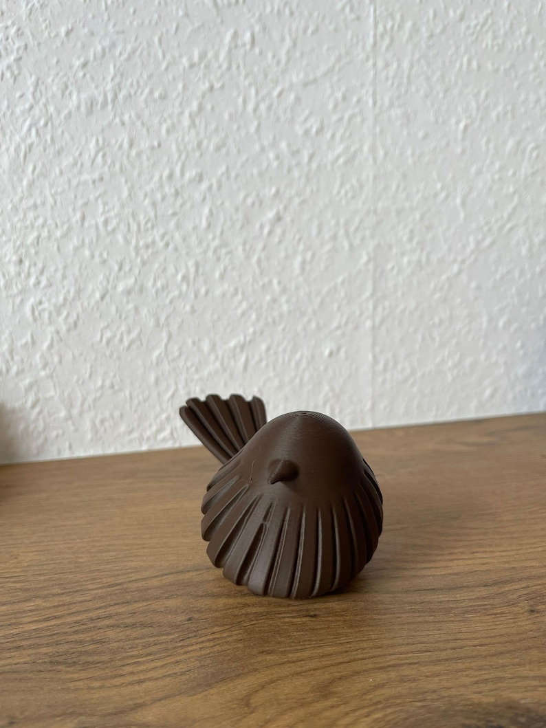 May include: A small, dark brown bird figurine with a rounded body and a textured, fan-shaped tail. The object features vertical ridges along its wings and a small, pointed beak, showcasing a smooth, matte finish throughout its entire structure.