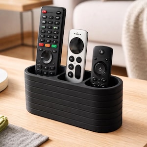 Peut inclure: Un organiseur de télécommandes noir et rainuré contenant trois télécommandes différentes. Les télécommandes sont noires et blanches. L'organisateur est posé sur une table en bois.