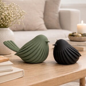 May include: Two bird figurines, one in matte olive green and one in matte black, featuring a stylized design with deep, curved grooves along the body and wings. The green bird is larger and positioned to the left, while the smaller black bird sits to the right, both facing toward the right side of the frame.