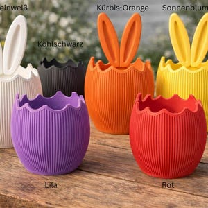 May include: A selection of vibrantly coloured, egg-shaped planters with bunny ear designs. The planters are in shades of white, black, orange, yellow, purple, and red. They feature a ribbed texture and are ideal for Easter decorations.