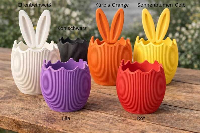 May include: A selection of vibrantly coloured, egg-shaped planters with bunny ear designs. The planters are in shades of white, black, orange, yellow, purple, and red. They feature a ribbed texture and are ideal for Easter decorations.