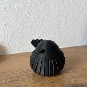 May include: A matte black bird figurine featuring a rounded body with vertical ribbed detailing along the sides and back. The bird has a small, pointed beak and a fanned tail, presented in a solid, monochromatic design with a smooth, uniform finish.