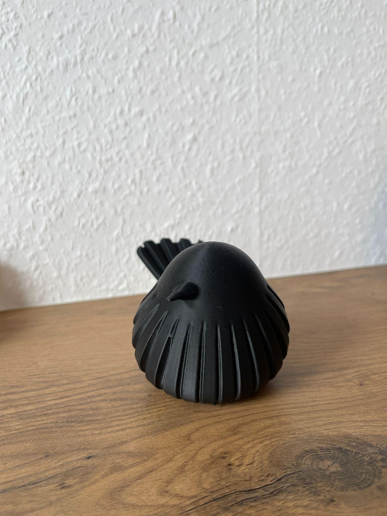 May include: A matte black bird figurine featuring a rounded body with vertical ribbed detailing along the sides and back. The bird has a small, pointed beak and a fanned tail, presented in a solid, monochromatic design with a smooth, uniform finish.