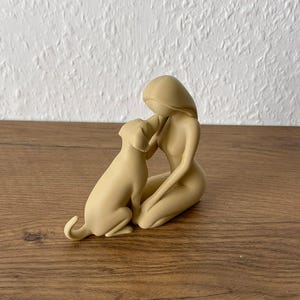 May include: A beige monochromatic figurine depicting a stylized human figure kneeling and interacting with a dog. The smooth, matte-finished sculpture shows the human figure leaning forward toward the dog, with both figures rendered in a simplified, abstract form.