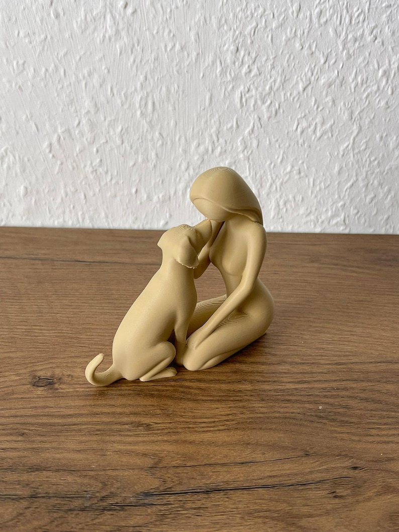 May include: A beige monochromatic figurine depicting a stylized human figure kneeling and interacting with a dog. The smooth, matte-finished sculpture shows the human figure leaning forward toward the dog, with both figures rendered in a simplified, abstract form.