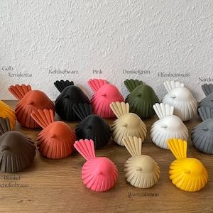 May include: A collection of small, stylized bird figurines in various colors including yellow, terracotta, black, pink, dark green, white, gray, dark chocolate brown, and desert tan. Each bird features a rounded body with vertical ridges.