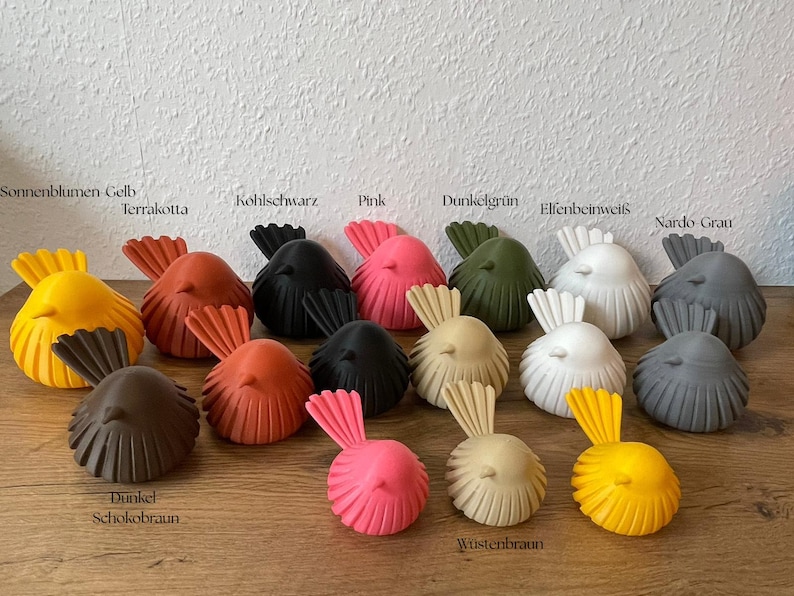 May include: A collection of small, stylized bird figurines in various colors including yellow, terracotta, black, pink, dark green, white, gray, dark chocolate brown, and desert tan. Each bird features a rounded body with vertical ridges.