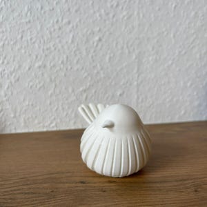 May include: A small, white, bird-shaped figurine with a smooth, rounded body and vertical, grooved detailing along its sides. The bird has a pointed beak and a fanned, textured tail, presented in a clean, solid white finish with a matte appearance.