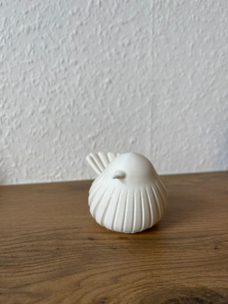 May include: A small, white, bird-shaped figurine with a smooth, rounded body and vertical, grooved detailing along its sides. The bird has a pointed beak and a fanned, textured tail, presented in a clean, solid white finish with a matte appearance.